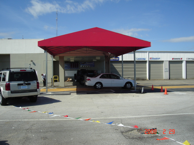 Red heavy-duty industrial awning providing shade for a commercial car service area, featuring a durable metal frame and open sides.